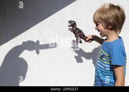 Kind mit Dinosaurier in der Hand und Silhouette des Mannes mit Schwert in der Hand vor Hintergrund der weißen Wand. Interessante kontrastierende Schatten. Ideen für Shad Stockfoto