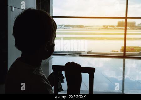 Silhouette eines kaukasischen Jungen mit Koffer, der am Fenster im Flughafen steht und Flugzeuge beobachtet, die auf den Flug warten. Alleinreisender-Konzept Stockfoto