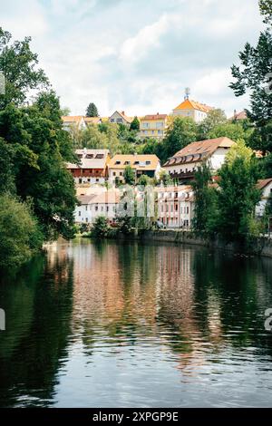 Eine ruhige Gemeinde am Fluss bietet charmante Häuser, die zwischen grünen Bäumen eingebettet sind. Das ruhige Wasser spiegelt die farbenfrohen Gebäude und die Umgebung wider Stockfoto