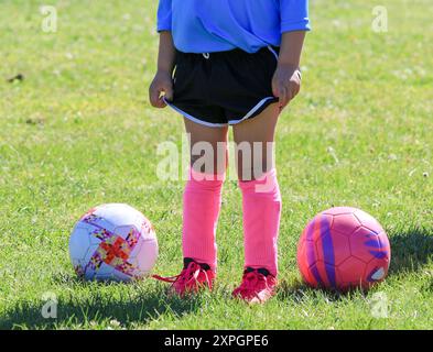 Ein junger Fußballspieler steht selbstbewusst auf einem grasbewachsenen Feld, trägt ein blaues Trikot, schwarze Shorts und knallrosa Socken. Zwei bunte Fußballbälle ruhen aus Stockfoto