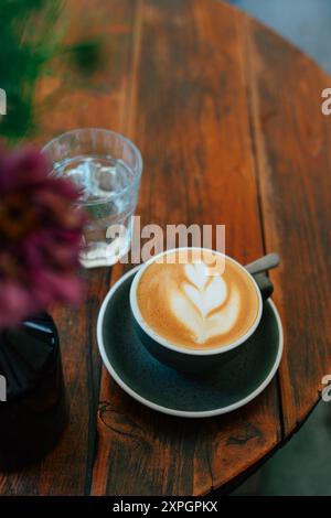 Eine frisch gebrühte Tasse Kaffee steht auf einem rustikalen Holztisch, der mit komplizierten Latte Art geschmückt ist. Daneben glitzert ein klares Glas Wasser im weichen Stockfoto