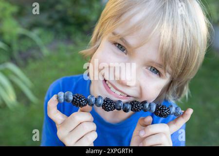 Das süße Kind hält Brombeeren und Heidelbeeren am Stock vor dem Gesicht. Fröhliches Kindergesicht mit saisonalen Sommerbeeren. Kinder, die Orga pflücken Stockfoto