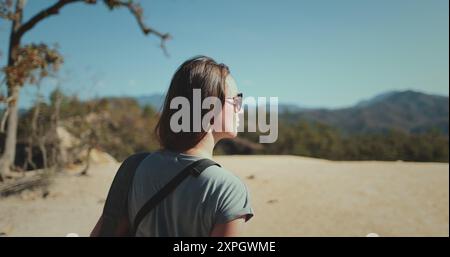 Eine junge Frau mit Sonnenbrille und blauem Hemd geht auf einem Sandweg auf einer tropischen Insel. Üppiges Laub und Berge sind in der Ferne sichtbar. Pai Canyon Chiang Mai, Thailand. Zeitlupe Stockfoto