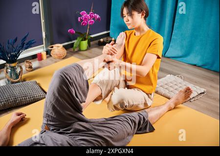 Masseurin, die thai-Yoga-Massage macht. Eine Therapeutin, die traditionelle Massagebehandlung durchführt und den Körper des männlichen Patienten dehnt. Der Kunde liegt auf der Matte und genießt eine Handmassage in ruhiger Atmosphäre. Stockfoto