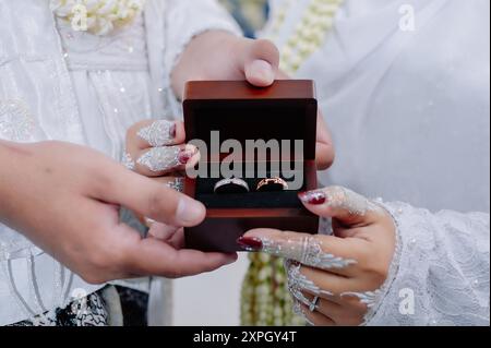 Die beiden asiatisch-indonesischen Bräutigame halten Eheringe in Holzkisten. Die Hände der Braut tragen Henna und Nagelschmuck Stockfoto