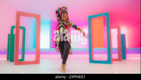 Porträt einer talentierten Black Female Künstlerin, die anmutige Bewegungen und ausdrucksvolle freudige Emotionen mit ihrer innovativen und energiegeladenen modernen Tanzroutine in einem farbenfrohen Neonlicht Studio zeigt. Stockfoto