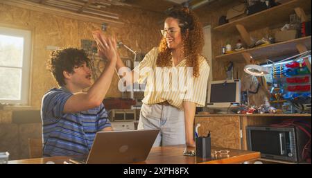 Kaukasischer männlicher Software-Engineer und hispanische weibliche Marketing-Direktorin sprechen und Laptop-Computer in der Garage zu Hause benutzen. Verschiedene Junge Startup-Gründer Arbeiten An Innovativem Online-Service. Stockfoto