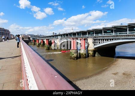 London - 06 10 2022: Mädchen schaut auf das Handy auf der Blackfriars Bridge und ein Touristenboot segelt unter der Blackfriars Rail Bridge. Stockfoto