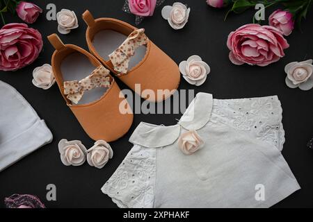 Babykleidung, Schuhe und Rosen auf schwarzem Hintergrund, Babyduschkonzept. Stockfoto