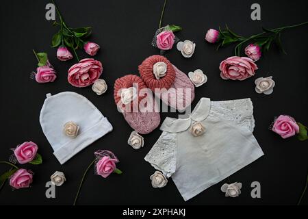 Babykleidung, Schuhe und Rosen auf schwarzem Hintergrund, Babyduschkonzept. Stockfoto