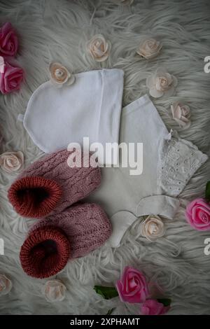 Rosafarbene Strickschuhe und ein weißer Hut auf weißem Fell, umgeben von rosa Rosen. Stockfoto