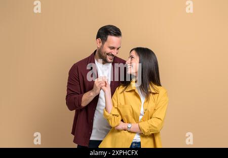 Romantischer junger Mann und Frau, die Hände halten und lächeln, während sie über beigefarbenem Hintergrund stehen. Attraktives Paar in lässigen Kleidern, das jeden einzelnen Blick ansieht Stockfoto