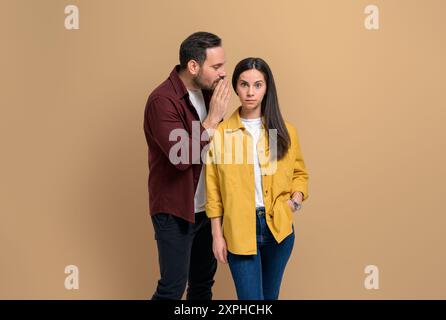 Schockierte Freundin mit erhöhten Augenbrauen, die zuhört, wie ihr Freund Geheimnisse in die Ohren flüstert. Ein junges Paar, das im Stehen über private Klatsch spricht Stockfoto