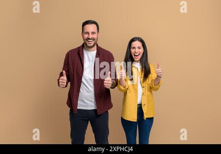 Charmante junge Frau und Mann in lässigen Kleidern zeigen Daumen hoch und lachen, während sie über beigefarbenem Hintergrund stehen. Fröhliches Paar, das in die Kamera schaut Stockfoto