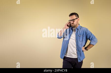Frustrierter und gestresster junger Geschäftsmann mit Hand in Hüfte, der über Smartphone isoliert vor gelbem Hintergrund diskutiert. Er trägt kluge Casuals Stockfoto