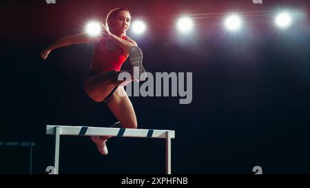 Weibliche Athletin in der Mitte der Luft, Sprung über die Hürde unter hellen Stadionlichtern, Agilität, Stärke und Entschlossenheit beim High-Energy Track and Field Event. Stockfoto