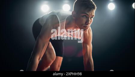 Starker junger Mann, der am Abend ein Rennen von der Startblockposition in einem dunklen Stadion startet. Porträt eines fitten männlichen Sprint Runners, der an einem internationalen Wettbewerb teilnimmt Stockfoto