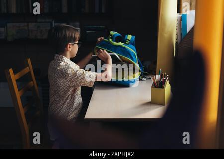 Der Junge, der sich auf die Schule vorbereitet, sitzt auf einem Stuhl und sammelt Schulsachen in seinem Rucksack Stockfoto