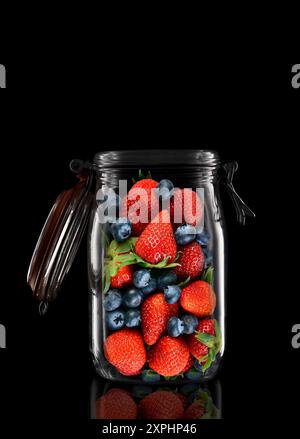 Erdbeeren und Heidelbeeren in einem Glasvorratsbehälter oder Dosenbecher isoliert auf Schwarz mit Reflexion, mit offenem Deckel. Stockfoto