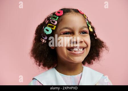 Ein junges afroamerikanisches Mädchen mit bunten Haarspangen lächelt hell vor einem rosa Hintergrund. Stockfoto