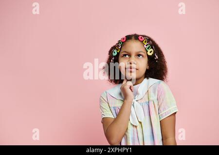 Ein schwarzes Mädchen mit farbenfrohen Haarspangen steht in einem pastellfarbenen Kleid vor rosa Hintergrund, und ihr nachdenklicher Blick ruft Wunder hervor. Stockfoto