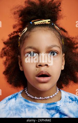 Ein schwarzes Mädchen mit lockigen Haaren, bunten Clips und blauem Outfit steht vor einem orangen Hintergrund. Stockfoto