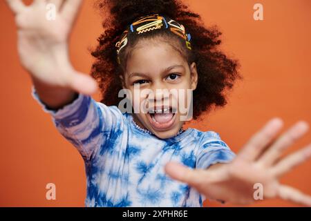 Ein junges afroamerikanisches Mädchen in einem blauen Krawattenhemd posiert mit ausgestreckten Händen vor einem orangefarbenen Hintergrund. Stockfoto