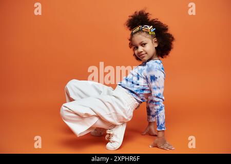 Bunte Haarspangen und trendiges Outfit auf afroamerikanischem Mädchen vor orangem Hintergrund. Blaues und weißes Hemd zum Binden mit weißer Hose. Stockfoto