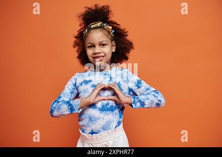 Ein junges afroamerikanisches Mädchen in einem trendigen Outfit steht vor einem leuchtenden orangen Hintergrund, lächelt und macht mit ihren Händen eine Herzform. Stockfoto