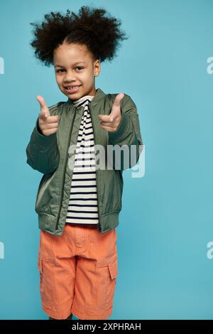 Ein bezauberndes schwarzes Mädchen in grüner Jacke, gestreiftem Hemd, orangen Shorts, Lächeln und Punkten mit beiden Händen vor hellblauem Hintergrund. Stockfoto