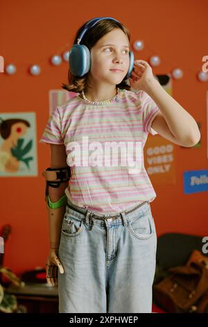 Vertikale mittellange Aufnahme eines fröhlichen Teenager-Mädchens mit Armprothese im y2k-Outfit, das auf dem Bett steht und Musik in den Kopfhörern hört Stockfoto