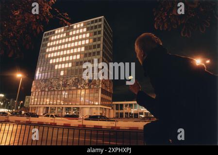 Ein Mann, der ein Handy hält, blickt auf eine herzförmige Lichtanzeige in einem Gebäude. Das beleuchtete Herz auf dem Wolkenkratzer symbolisiert Liebe und Verbundenheit und kontrastiert urbanes Nachtleben mit herzlichen Emotionen. Stockfoto