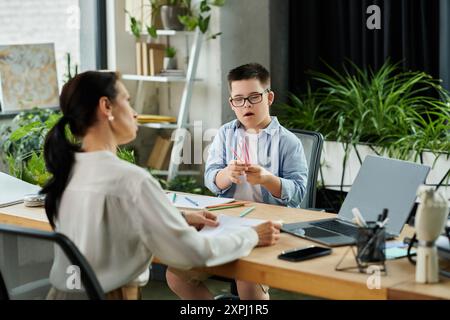 Eine Mutter arbeitet an ihrem Laptop, während ihr Sohn mit Down-Syndrom am Tisch neben ihr mit Buntstiften spielt. Stockfoto