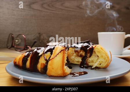 Teller mit leckerem SchokoladenCroissant und einer Tasse dampfenden Kaffee im Hintergrund Stockfoto