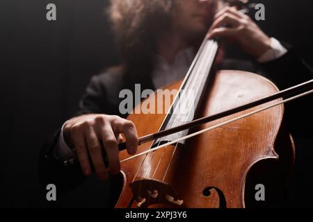 Beschnittene Aufnahme eines nicht erkennbaren, talentierten männlichen Cello-Spielers, der klassische Musik in dunklem Studio mit dunklem Licht aufführt, Fokus auf Violoncello-Bogen Stockfoto