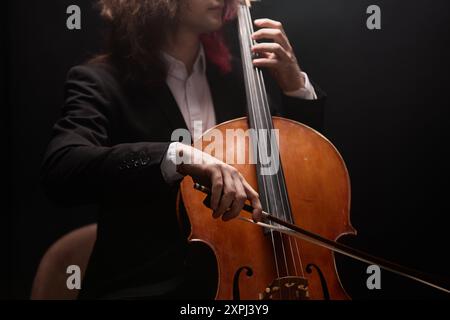 Beschnittene Aufnahme eines nicht erkennbaren männlichen Musikers mit langen Haaren, der Cello spielt und klassische Musik auf schwarzem Hintergrund in einem dämmerlichen Aufnahmestudio aufführt Stockfoto