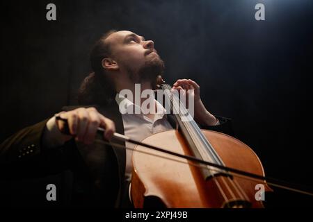 Niedriger Blickwinkel eines bewegten männlichen Musikers, der nachdenklich nach oben blickt, während er Cello in einem dunklen Studio mit Oberlicht und Kopierraum spielt Stockfoto