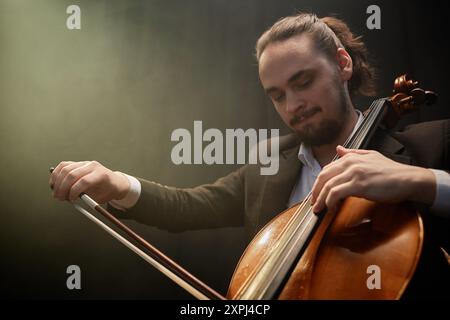 Porträt eines konzentrierten männlichen Musikers in schwarzem Anzug, der anmutig Cello spielt, in dunklem Studio mit Oberlicht, Kopierraum Stockfoto