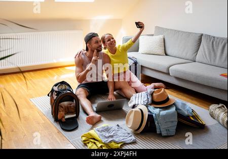 Ein junges Paar, das zu Hause auf dem Boden sitzt und ein Selfie macht, während es seine Reisetaschen packt. Stockfoto