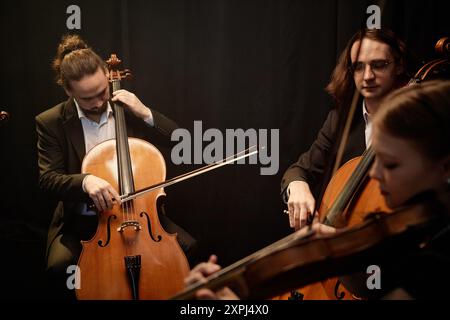 Drei sinfonische Orchestermusiker spielen Celli und Violinen, während sie im Kreis sitzen und klassische Musik vor schwarzen Vorhängen im Kontrastlicht spielen Stockfoto