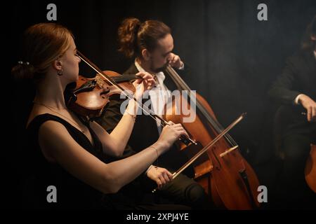 Seitenansicht von Kammermusikerinnen und -Musikern, die mit Leidenschaft Geige und Cello spielen, während eines Konzerts mit sinfonischen Quartetten auf der Bühne mit schwarzen Vorhängen Stockfoto