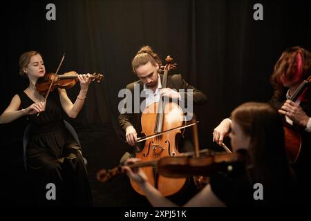 Männlicher klassischer Künstler, der leidenschaftlich Cello spielt, mit Streichquartettmusikern, die im Atelier im Kreis sitzen, mit Kontrastlicht gegen schwarze Vorhänge Stockfoto