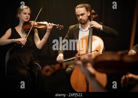 Virtuose weibliche und männliche Streicher spielen Violine und Cello auf der Bühne mit Kontrastlicht gegen schwarze Vorhänge Stockfoto