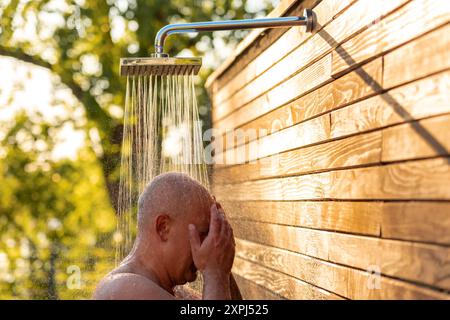 Mann unter Außendusche bei Sonnenuntergang. Konzept der Entspannung und erfrischenden Abendroutine Stockfoto