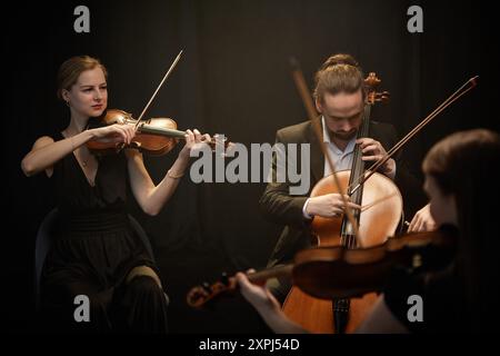 Professionelle weibliche und männliche Streicher spielen Violine und Cello mit Kammerquartett vor schwarzen Vorhängen Stockfoto