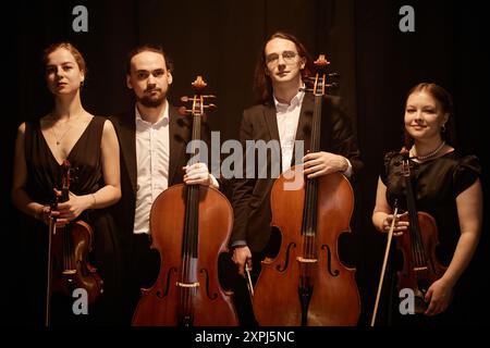 Gruppenporträt des Klassikers des Streichquartetts mit Blick auf die Kamera, während Sie auf der Bühne stehen, mit warmem Oberlicht vor schwarzen Vorhängen Stockfoto