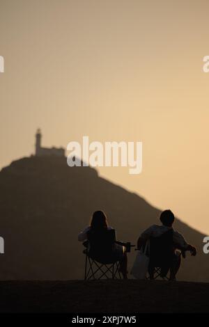 Ein Paar sitzt auf einem Hügel und beobachtet den Sonnenuntergang. Die Frau trägt ein weißes Hemd und der Mann trägt eine schwarze Jacke. Die Szene ist Peacefu Stockfoto