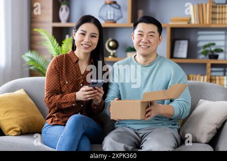 Fröhliches asiatisches Paar genießt den Moment, zusammen ein neues Paket zu eröffnen. Frau, die ein Smartphone hält, während der Mann die Lieferung auspackt. Fröhliche und entspannte Atmosphäre im gemütlichen Wohnzimmer. Stockfoto