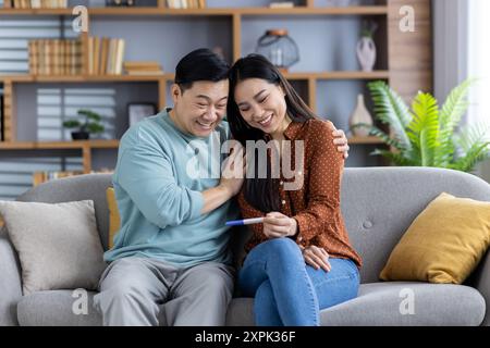 Asiatisches Paar feiert positive Schwangerschaftstestergebnisse zu Hause. Glückliche zukünftige Eltern genießen einen Moment der Freude und Vorfreude, während sie auf dem Sofa sitzen. Ich erwarte ein neues Baby und war fröhlich. Stockfoto