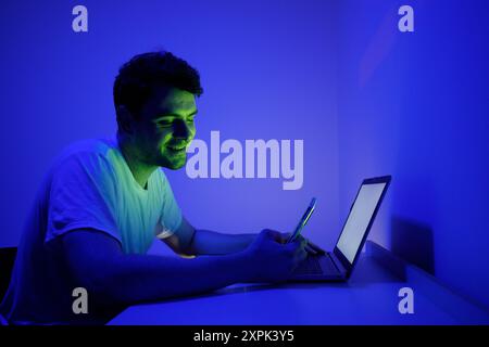 Ein junger Mann sitzt an einem Schreibtisch, beleuchtet durch ein sanftes blaues Licht, fokussiert auf seinen Laptop, während er ein Smartphone in einer Hand hält. Die Umgebungsbeleuchtung wird erzeugt Stockfoto